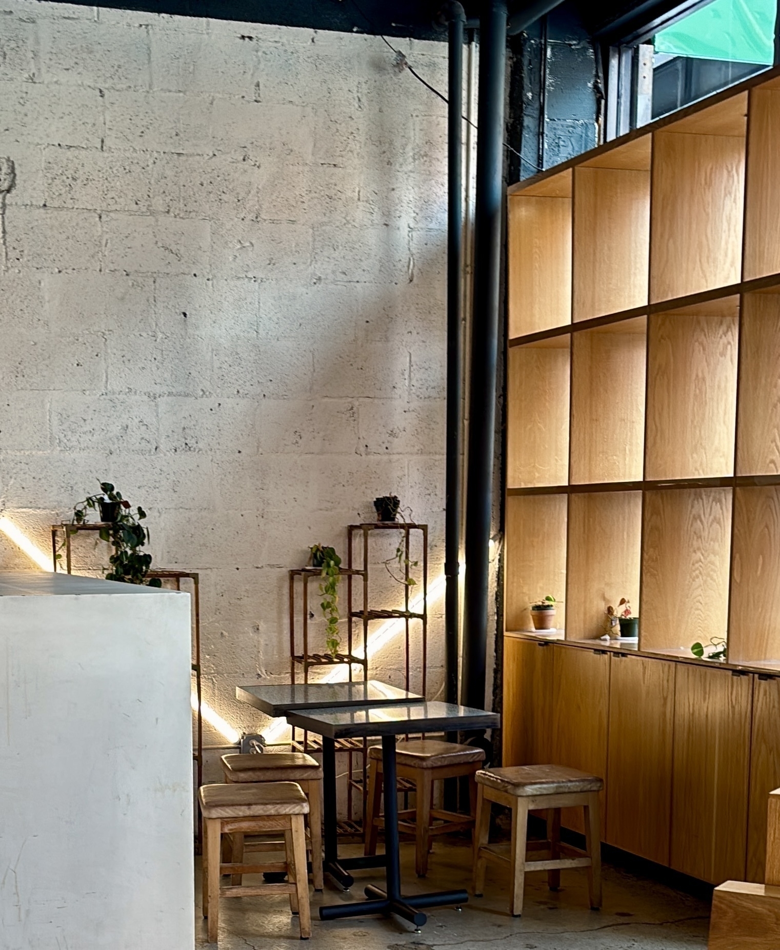 A cozy, minimalist café corner features wooden shelves, small plants, and a simple table with stools.