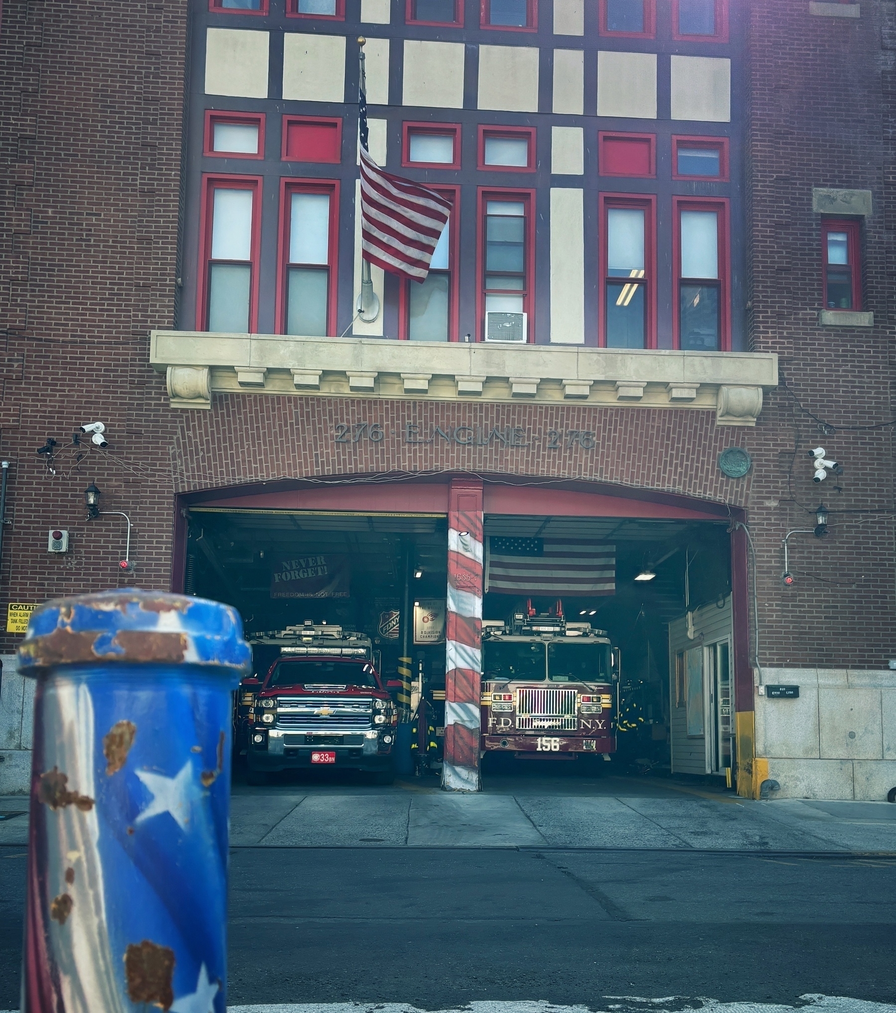 A fire station with an American flag and two fire trucks parked inside is visible behind a blue and white star-spangled bollard.