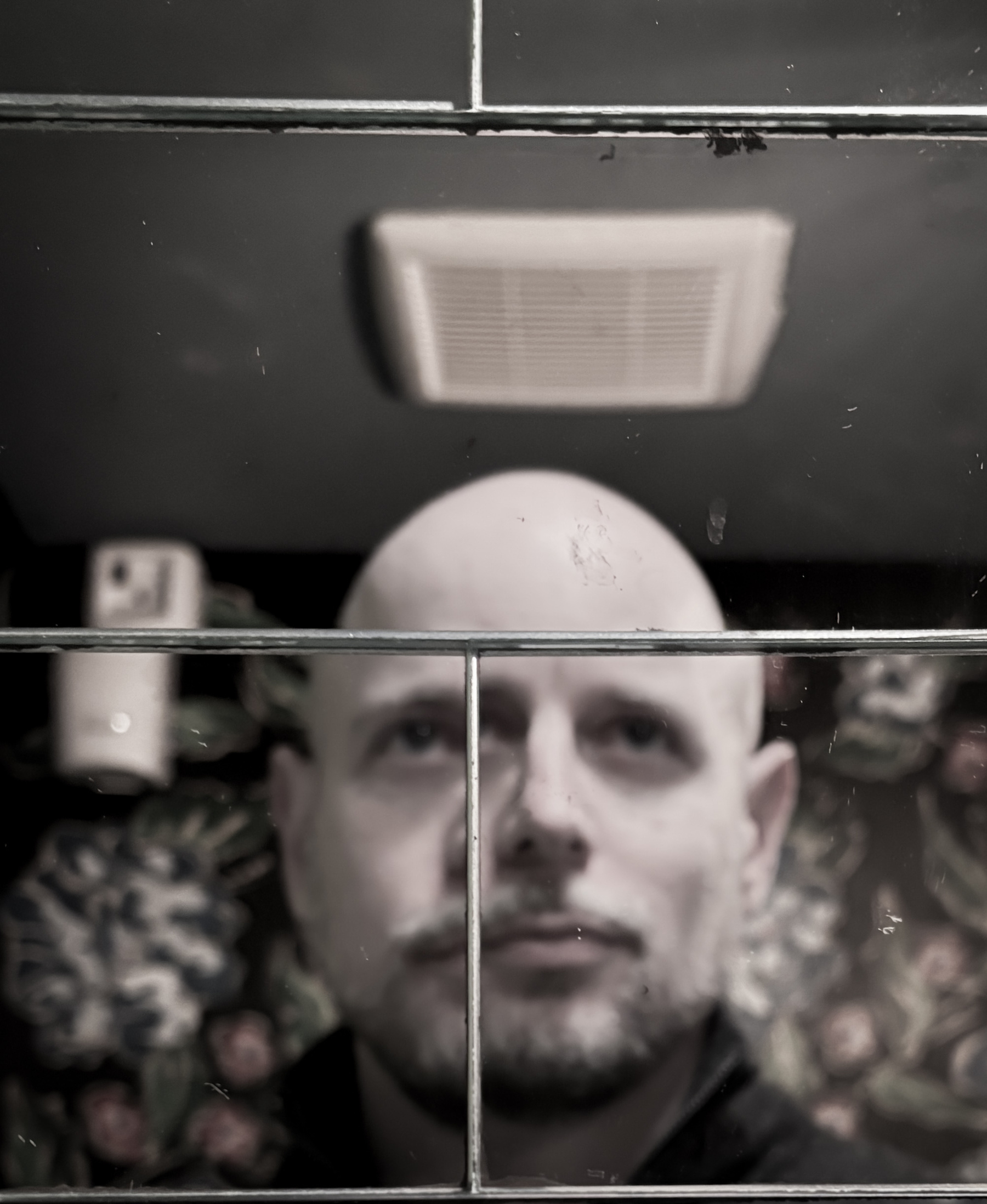 Auto-generated description: A bald man is reflected in a tiled bathroom mirror with a floral-patterned wall in the background.