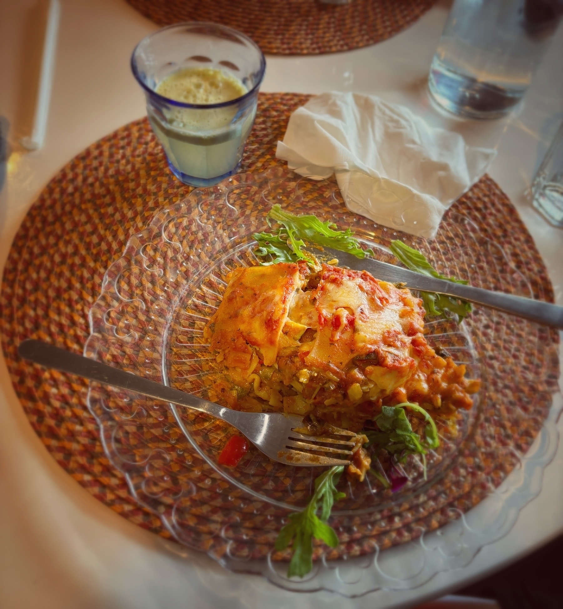 A plate of lasagna with a side of greens is set on a glass plate on top of a woven placemat, accompanied by a drink and a napkin.