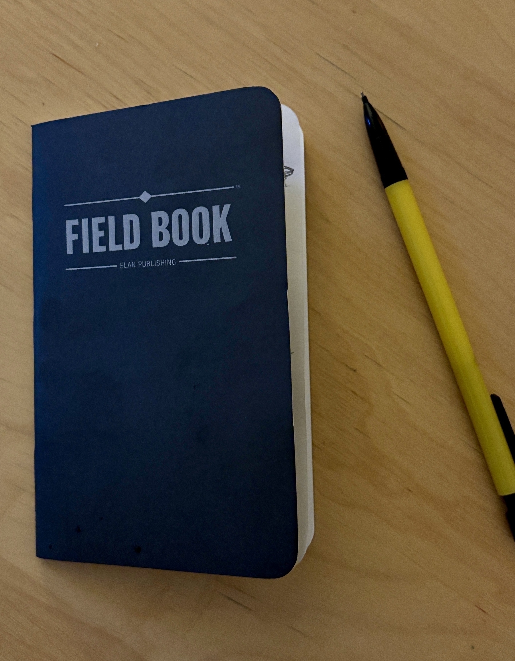 A black field notebook lies on a wooden table next to a yellow and black pen.