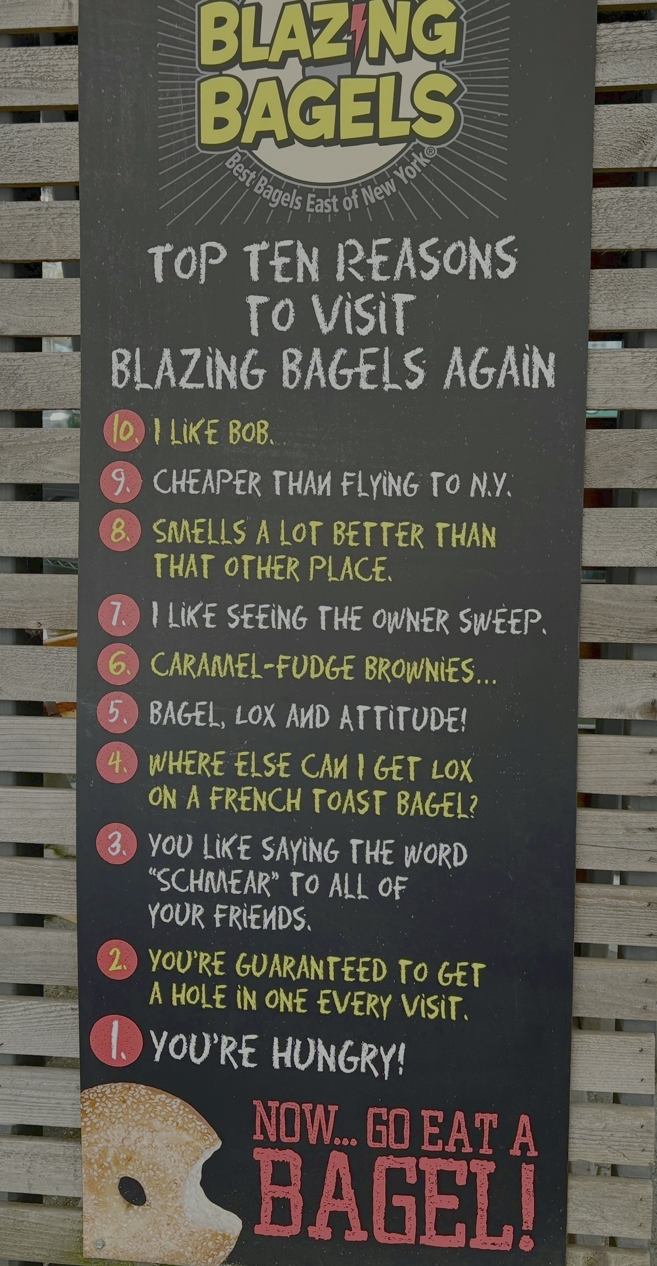 A sign for Blazing Bagels lists humorous reasons to visit, such as liking Bob, enjoying caramel-fudge brownies, and the guarantee of getting a hole in every visit.