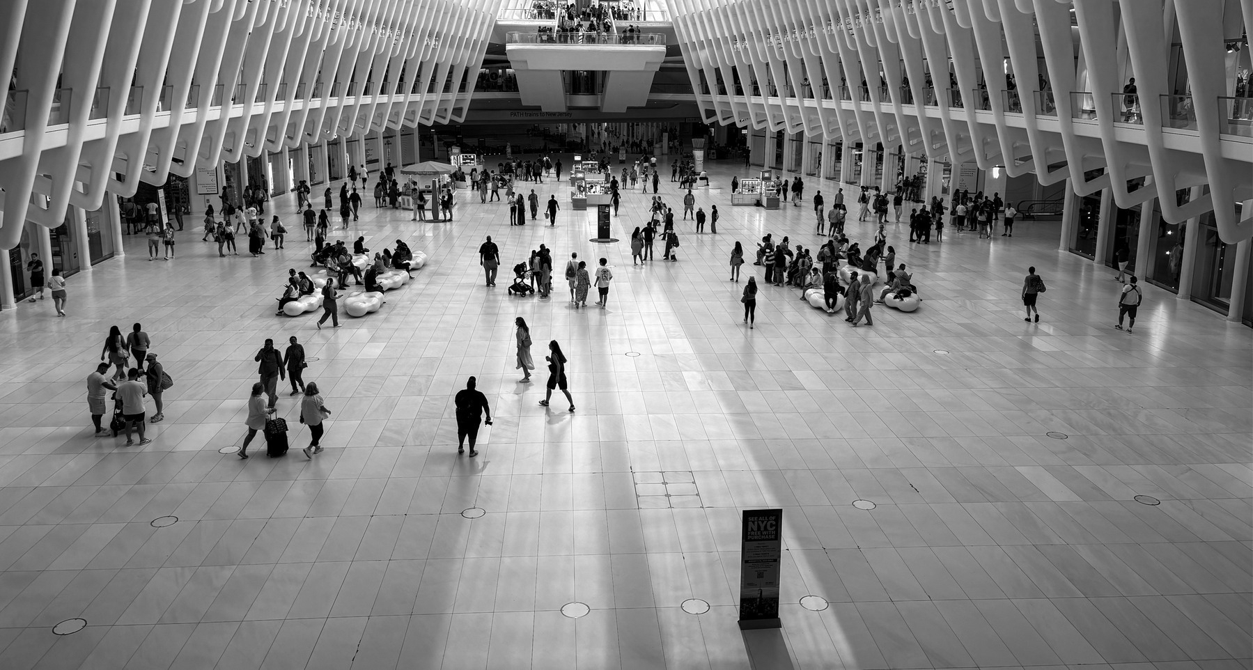 The inside of The Oculus World Trade Center Transportation Hub: A spacious indoor area with a high ceiling, large windows, and numerous people walking and sitting on benches. It looks like the inside of a giant dinosaur skeleton.