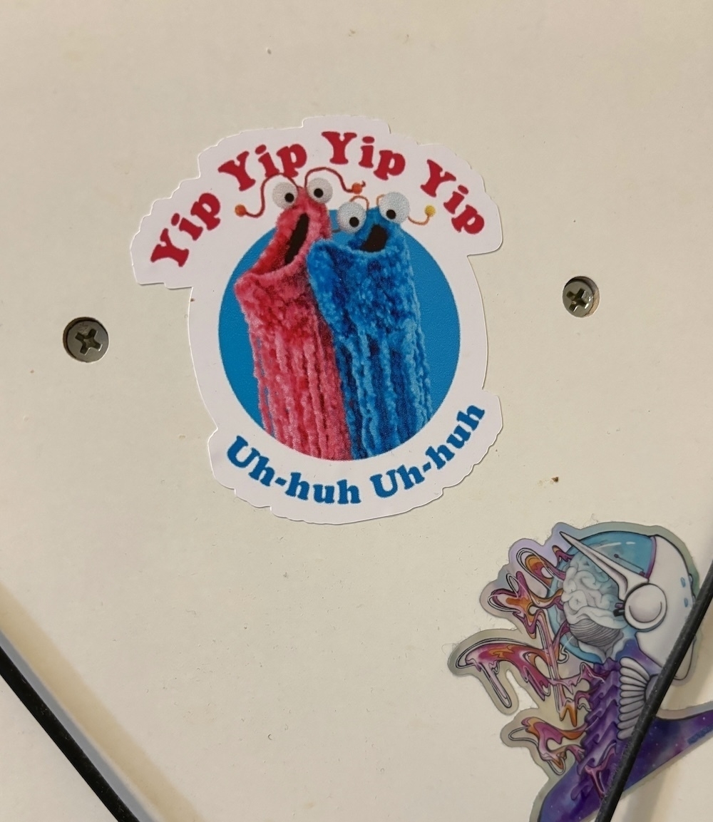 Auto-generated description: A sticker featuring two alien-like characters with googly eyes, one red and one blue, with text around them and another sticker of a colorful, artistic character nearby.