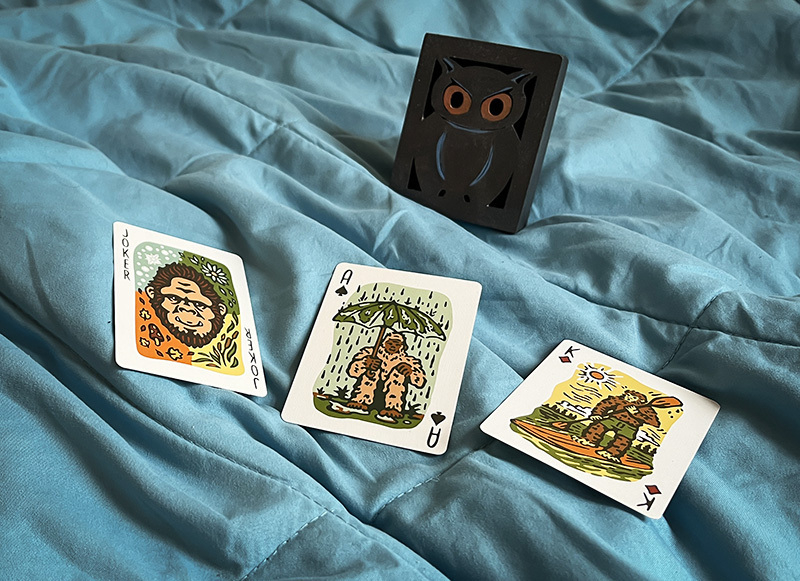 Three cards laying on a blanket: a Joke, an Ace and a King. They illustrate monkeys. A wooden box with an owl curved into it.