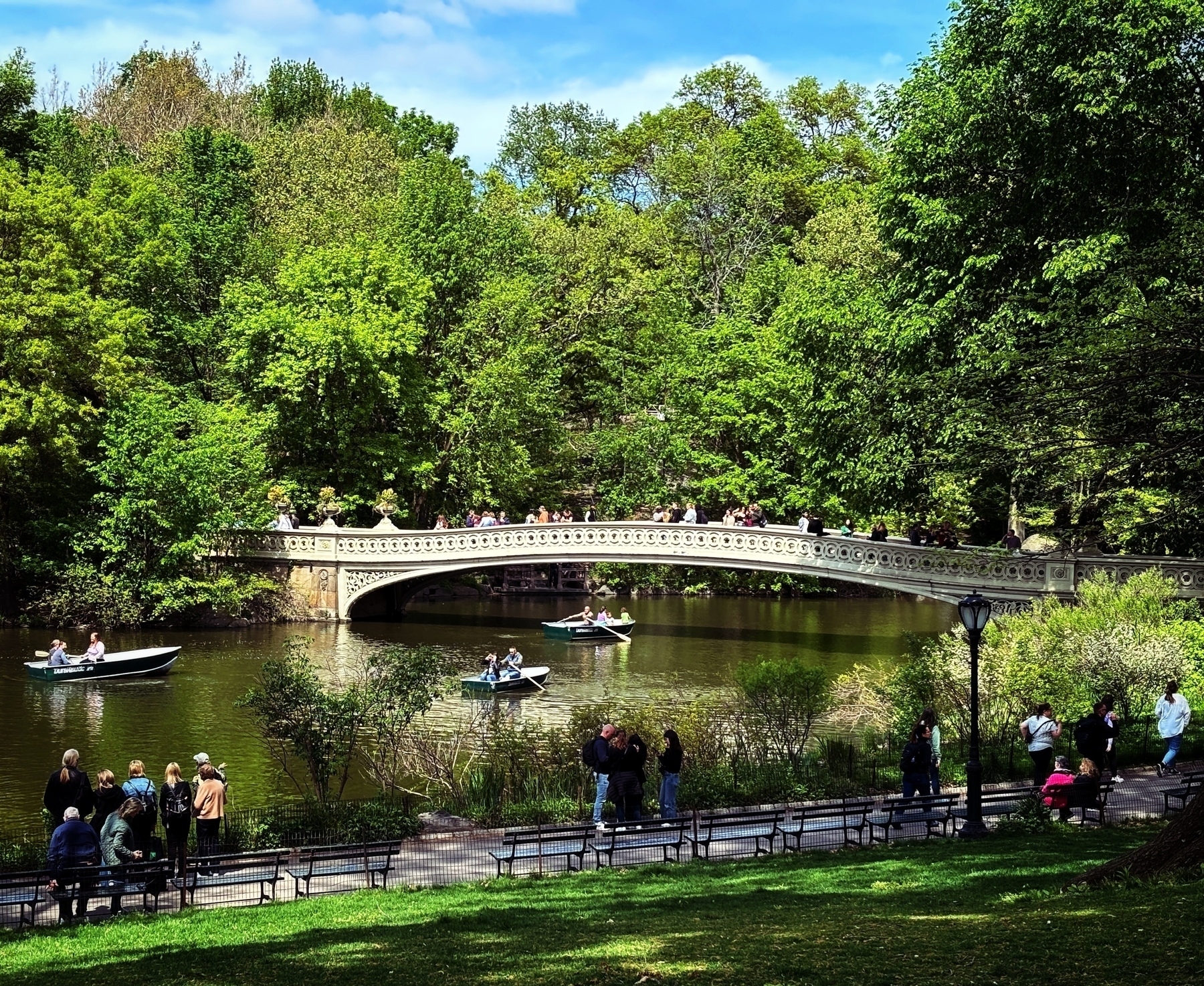 I wooden bridge over a calm brook in Central Park. Green trees and grass, people walking about, beautiful spring day