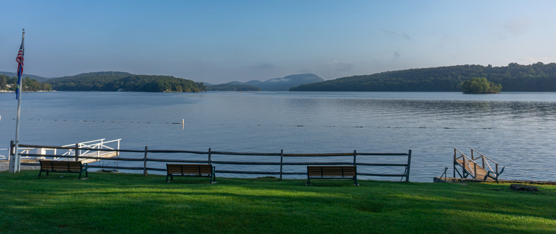 Auto-generated description: A serene lakeside view features lush green grass, benches, a dock, and distant hills under a blue sky.