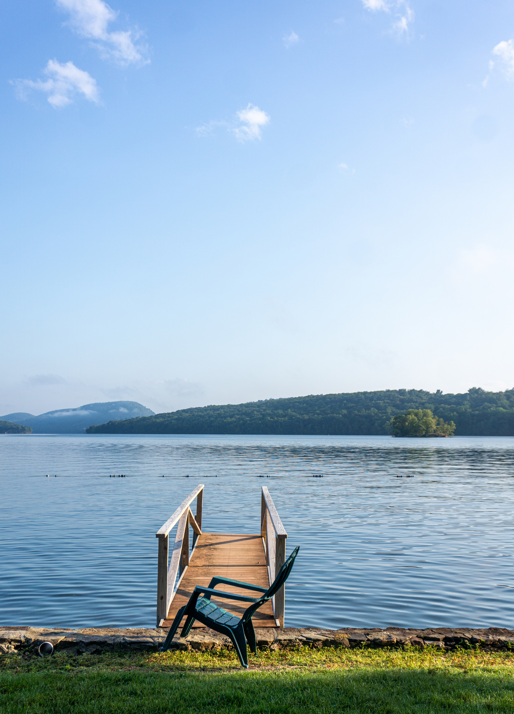 Auto-generated description: A small dock with a chair in the foreground overlooks a calm lake surrounded by gently sloping hills.