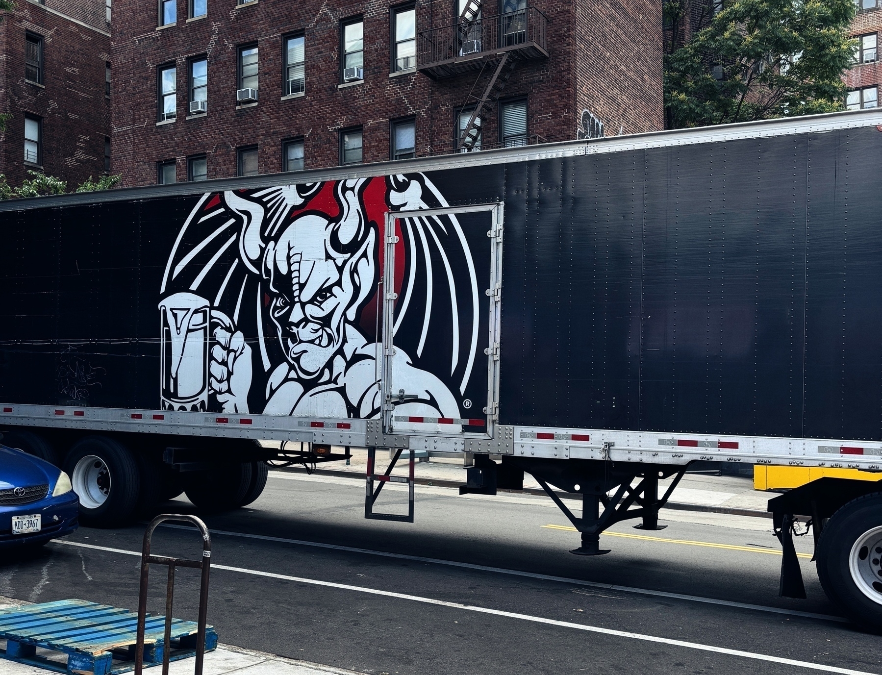 A large black trailer features a graphic of a menacing creature with wings and horns holding a mug against a backdrop of an urban street with brick buildings.&10;