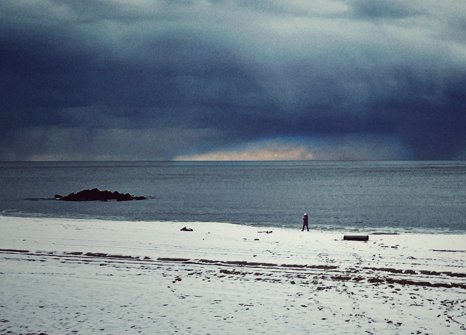 Snow on the beach, a person walking against the sea in the background. Dark blue clouds are hanging above