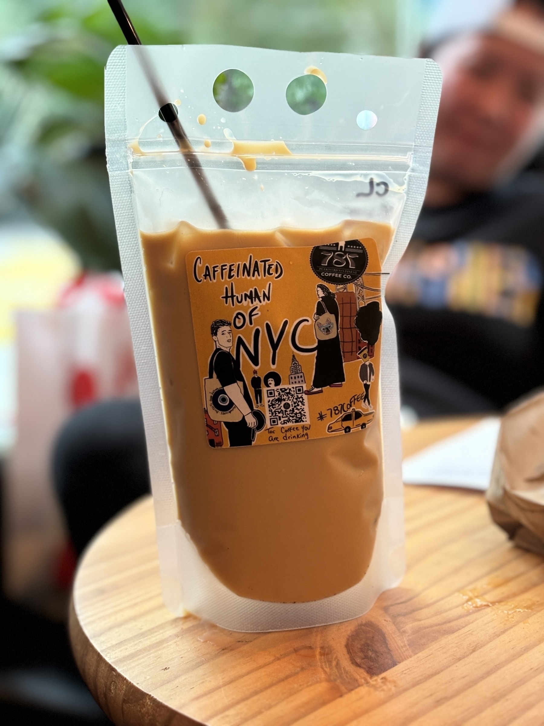 Auto-generated description: A beverage in a pouch labeled Caffeinated Human of NYC is placed on a wooden table with a straw inserted.