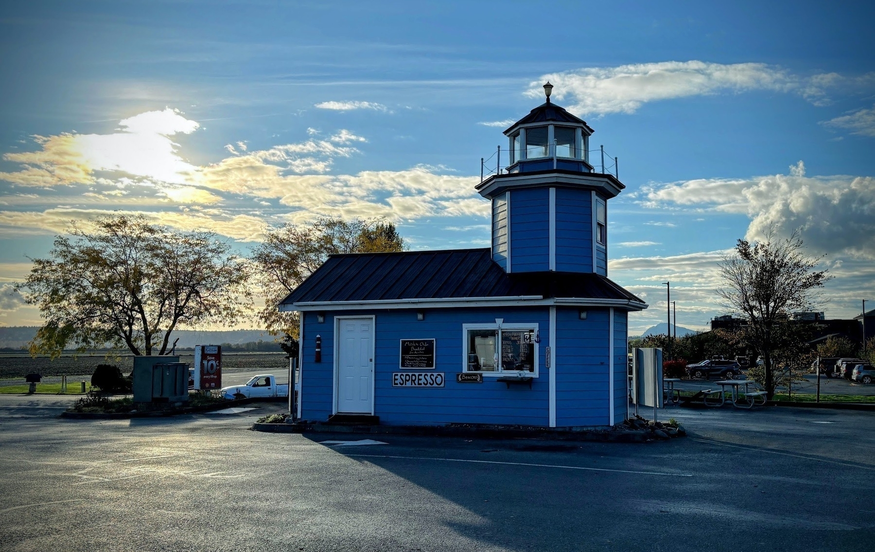 A small, blue lighthouse-shaped building serves as an espresso stand in a parking lot of a gas station