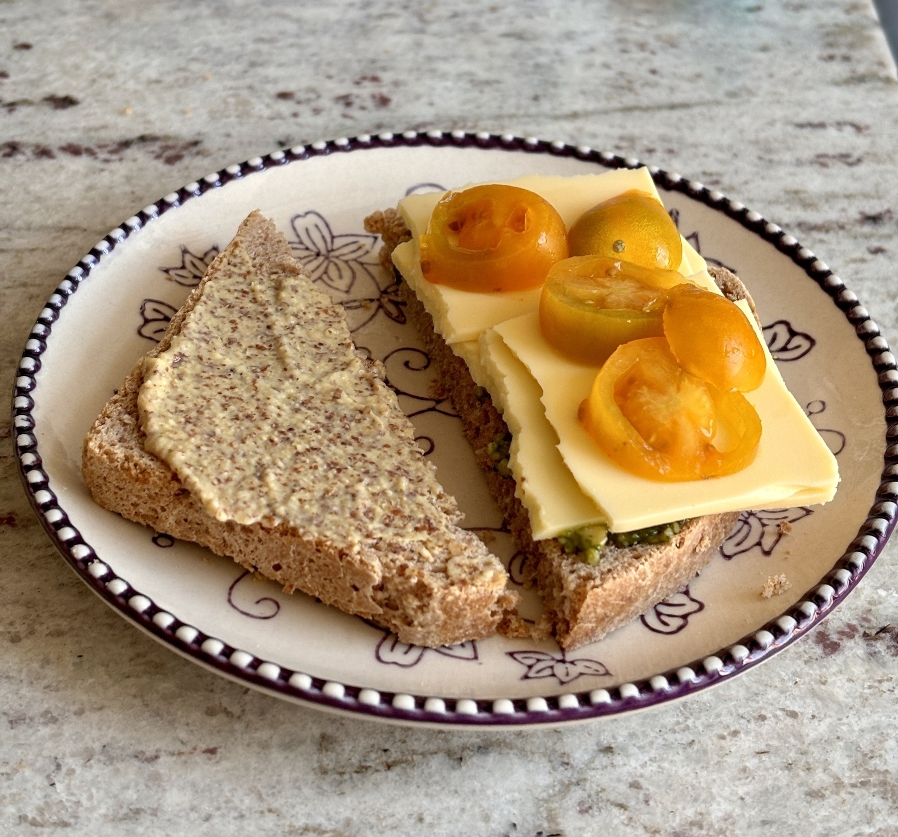 Auto-generated description: A plate holds a triangular sandwich with one slice of cheese and cut yellow cherry tomatoes on top of a bread slice spread with mustard.