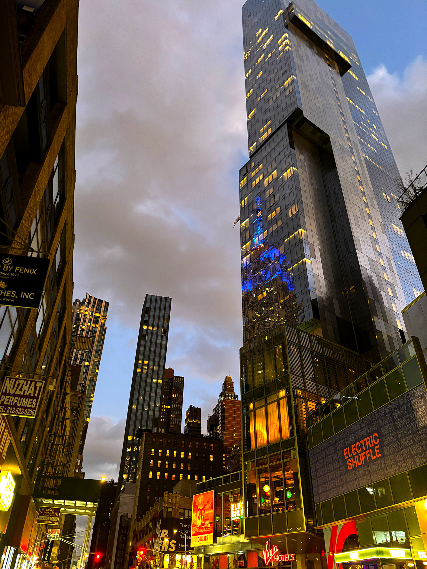 Auto-generated description: A tall, modern skyscraper towers over a bustling city street at dusk, with glowing signs and vibrant lights.