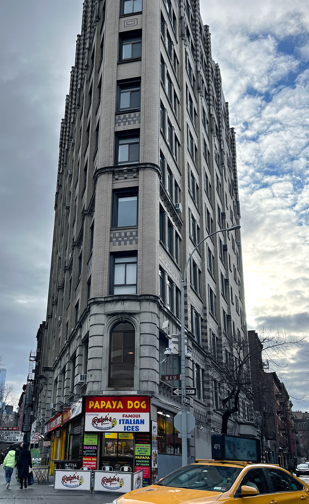 A tall building, trapozoid shape. its sharp corner faces the viewer, with a single column of windows going up. There are city streets on both sides of the building, with a yellow cab in front of the building.