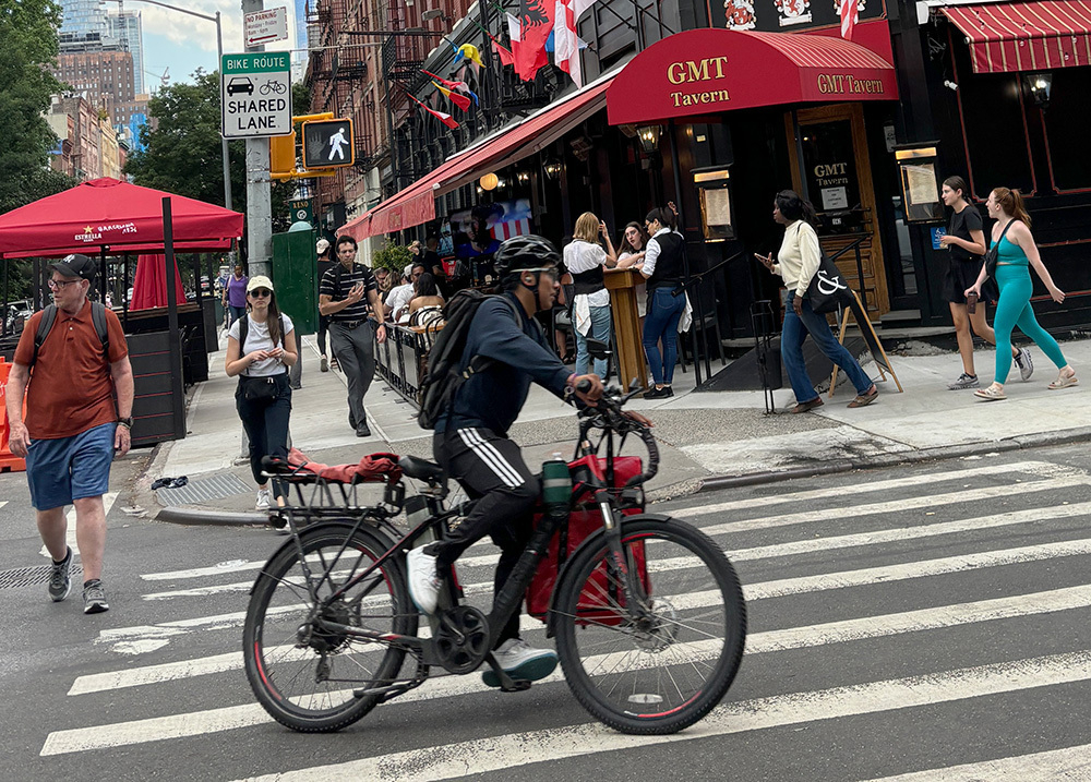 A cyclist rides through in a bustling new york city street with pedestrians and patrons walk outside a tavern on a street corner.