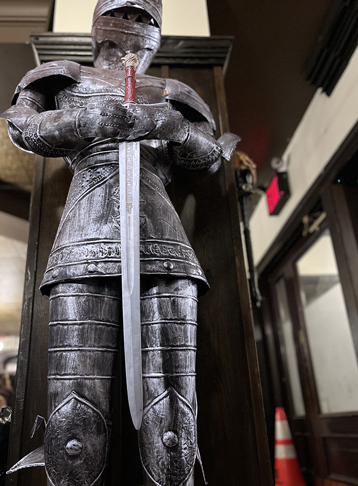 A suit of armor, a knight, holding a sord standing tall. An exit sign in the background with an orange cone.