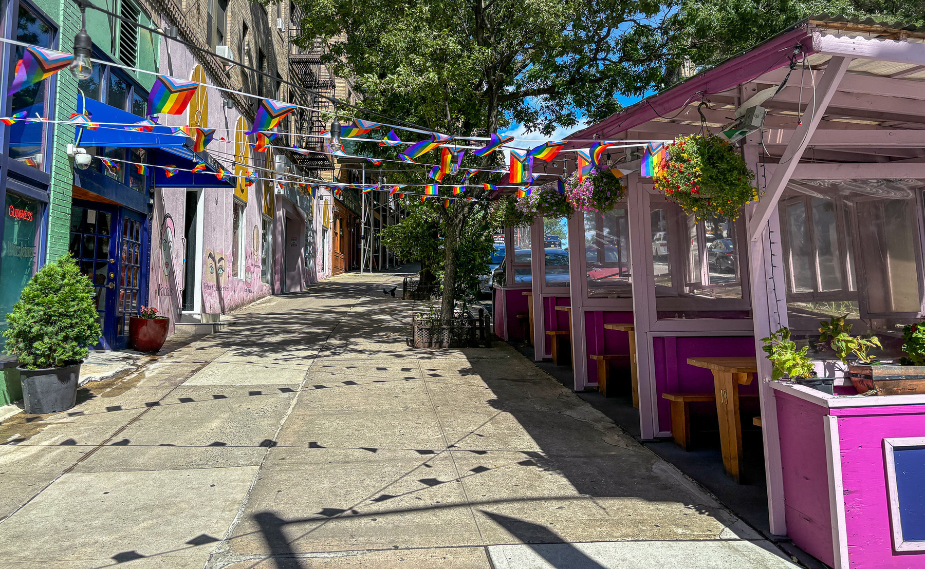 A colorful outdoor cafe with rainbow flags and vibrant decorations lines a pedestrian walkway under a clear blue sky.