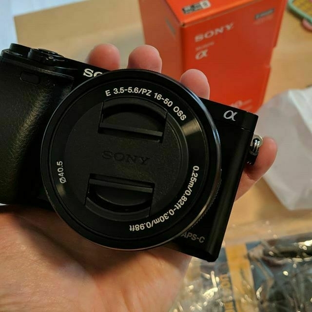 Auto-generated description: A person is holding a Sony Alpha mirrorless camera with its lens cap on, next to its packaging box.