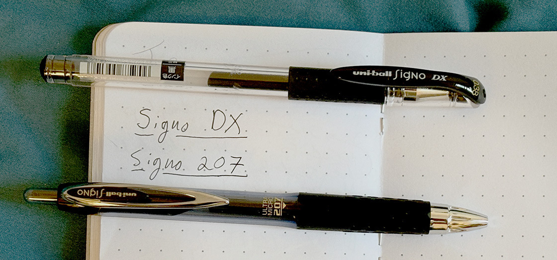 Auto-generated description: Two pens, labeled Signo DX and Signo 207, are placed on an open notebook with dotted pages.