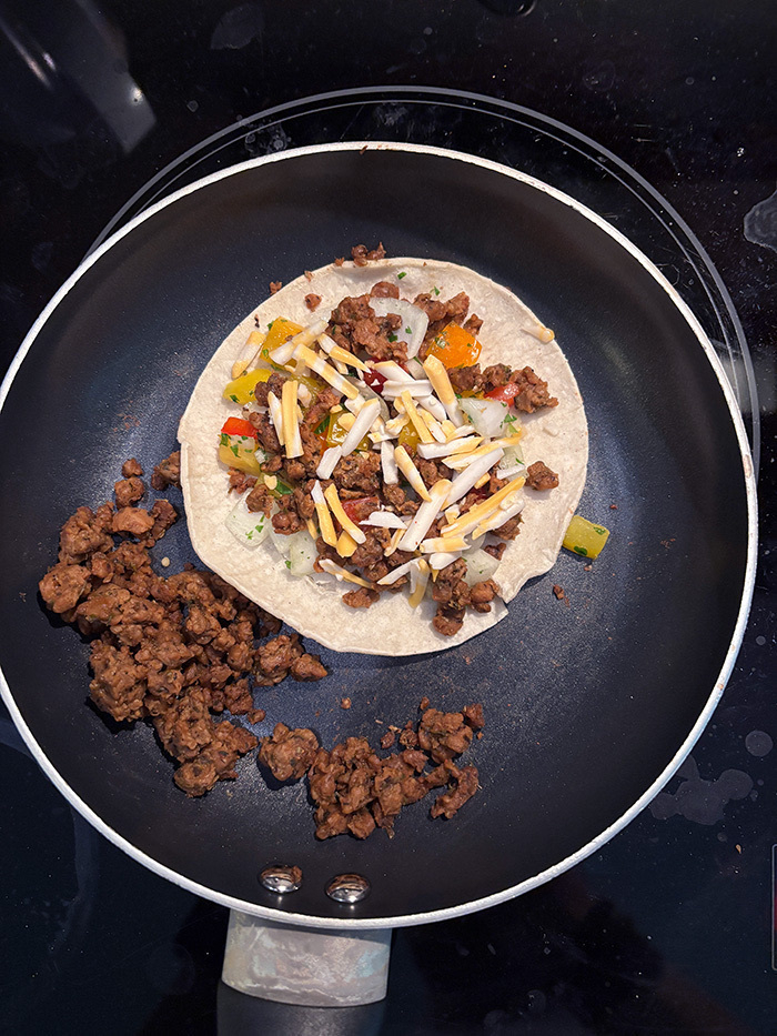 A tortilla is being cooked in a pan, topped with vegan ground meat (chorizo), diced vegetables (pico de gallo), and shredded vegan cheese.