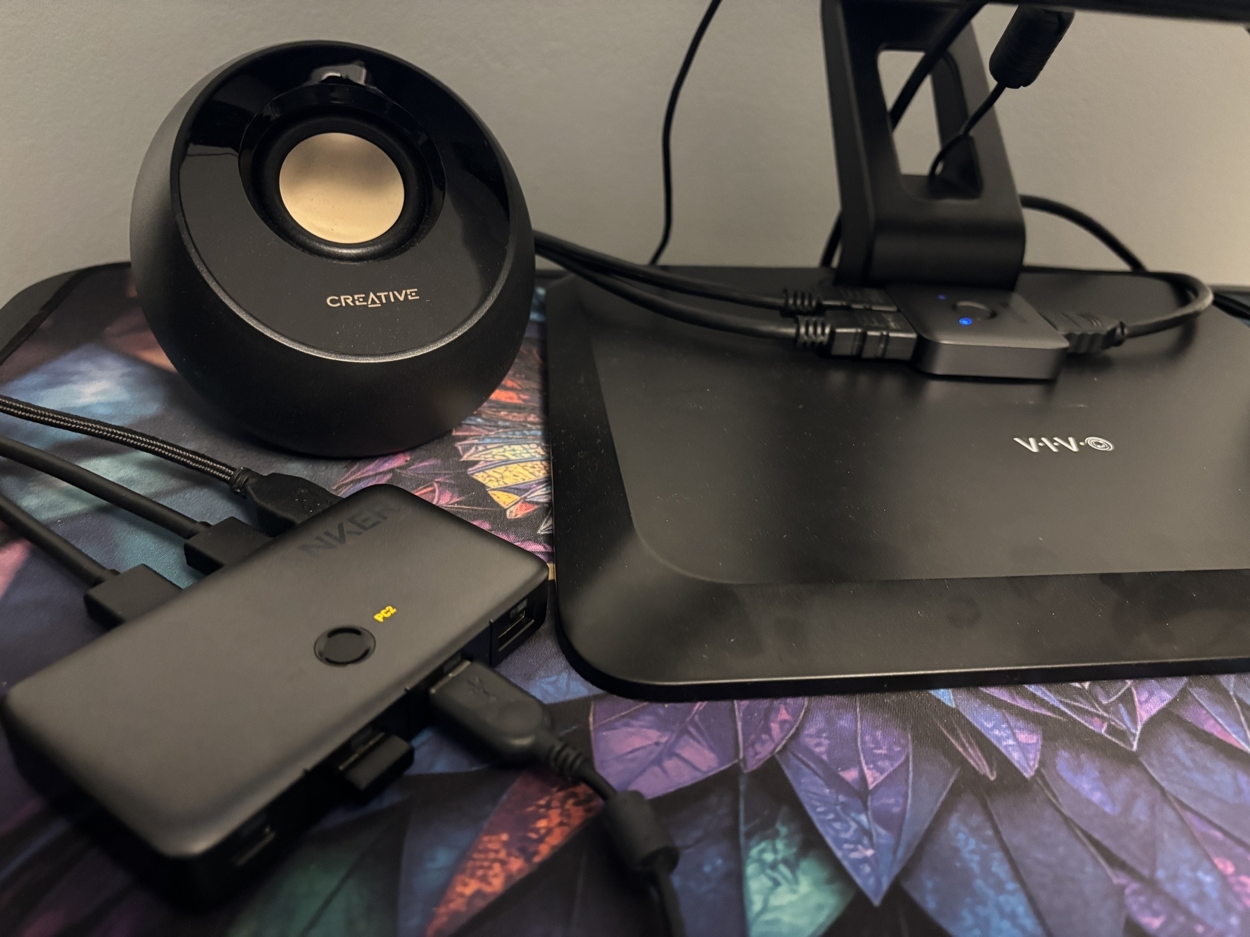 A desktop setup with a monitor stand, a speaker, and a connected electronic device (USB switch) on a colorful patterned surface.