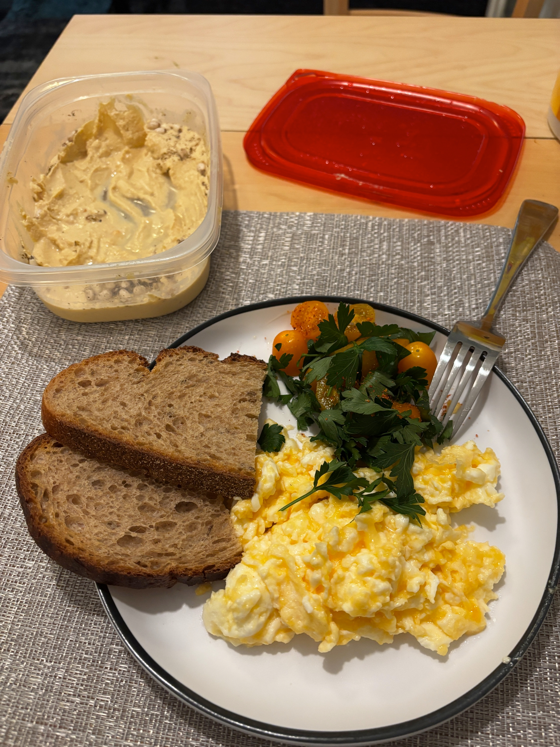 Auto-generated description: A breakfast plate features scrambled eggs garnished with parsley, cherry tomatoes, and slices of brown bread, with a side of hummus in a container.