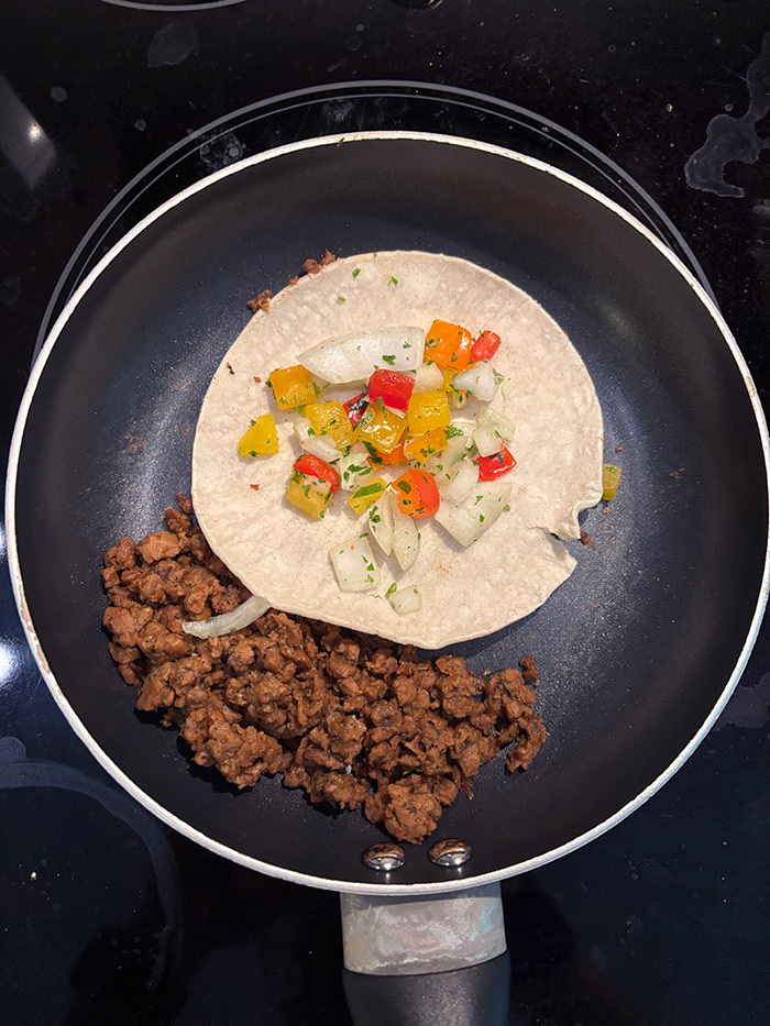 A tortilla topped with diced vegetables (pico de gallo) is placed in a pan alongside vegan ground meat (chorizo).