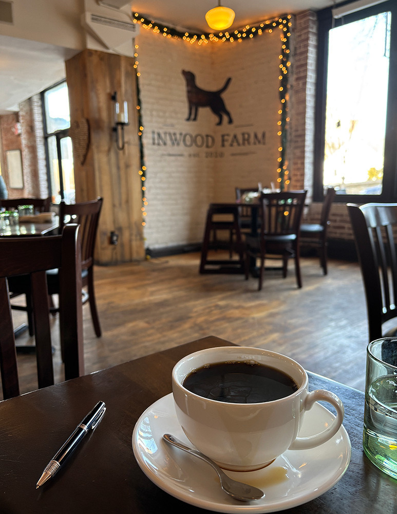 Auto-generated description: A cozy cafe features a cup of coffee on a wooden table with a pen, a glass of water, and a wall decorated with a silhouette of a dog and the words INWOOD FARM surrounded by string lights.