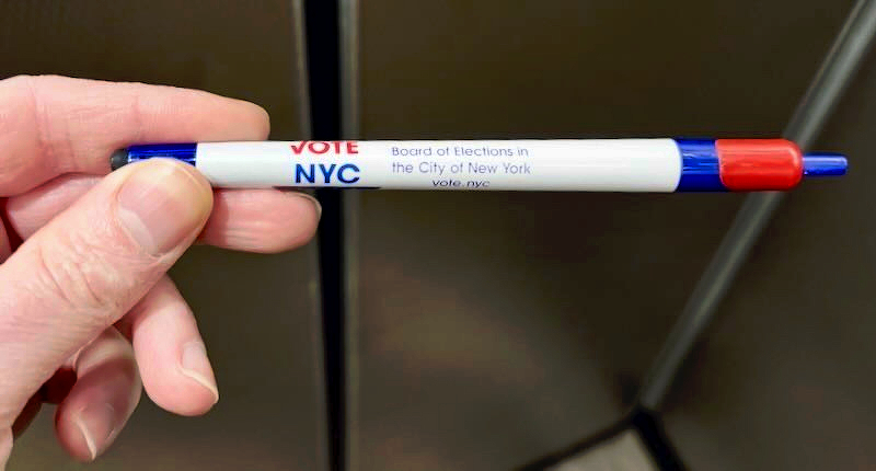 A hand is holding a red, white, and blue pen with VOTE NYC and election-related text on it.