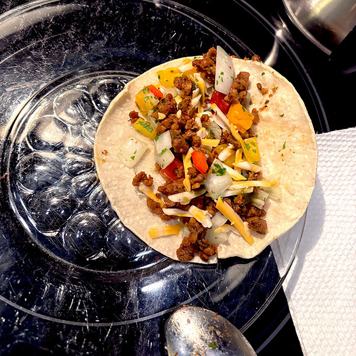 A taco filled with vegan ground meat (chorizo), diced vegetables (pico de gallo), and vegan cheese is placed on a clear, glass textured plate beside a spoon and napkin.