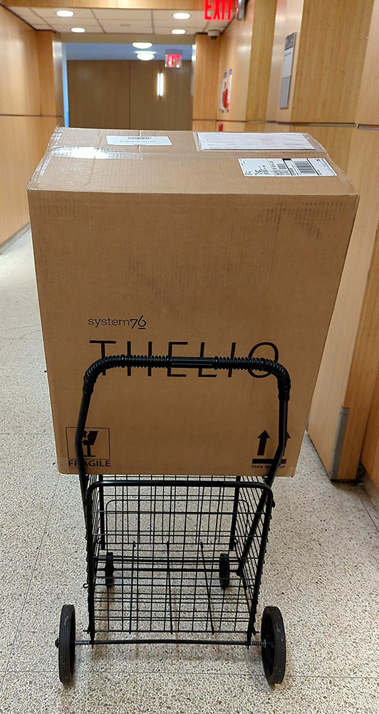 Auto-generated description: A large cardboard box labeled system76 is placed on a wire cart in a hallway.