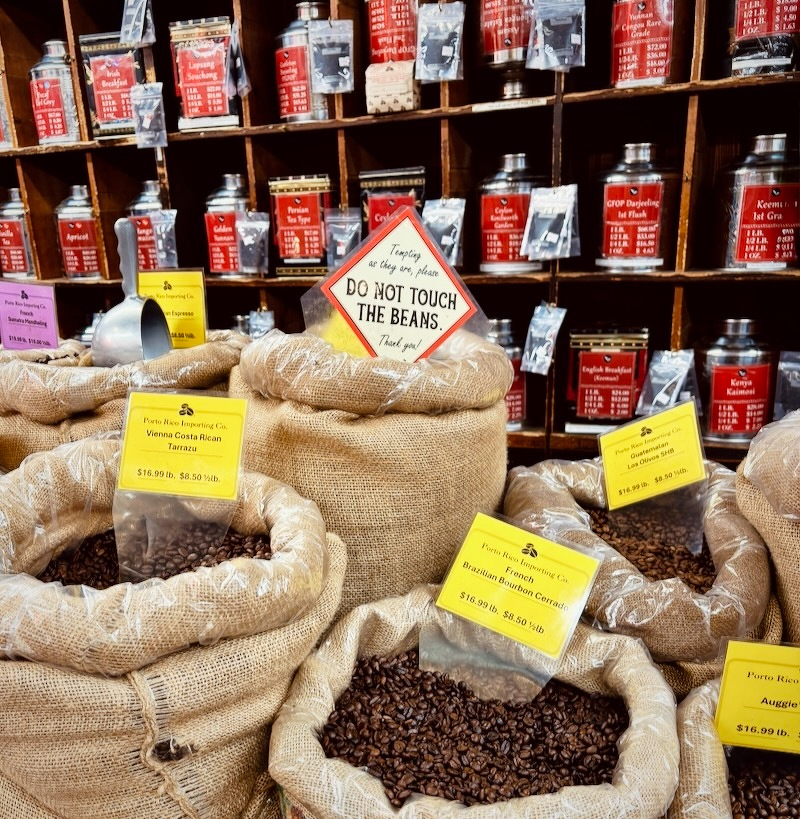 Auto-generated description: Bags of coffee beans are displayed with signs and price tags in front of a shelf filled with jars of various products.