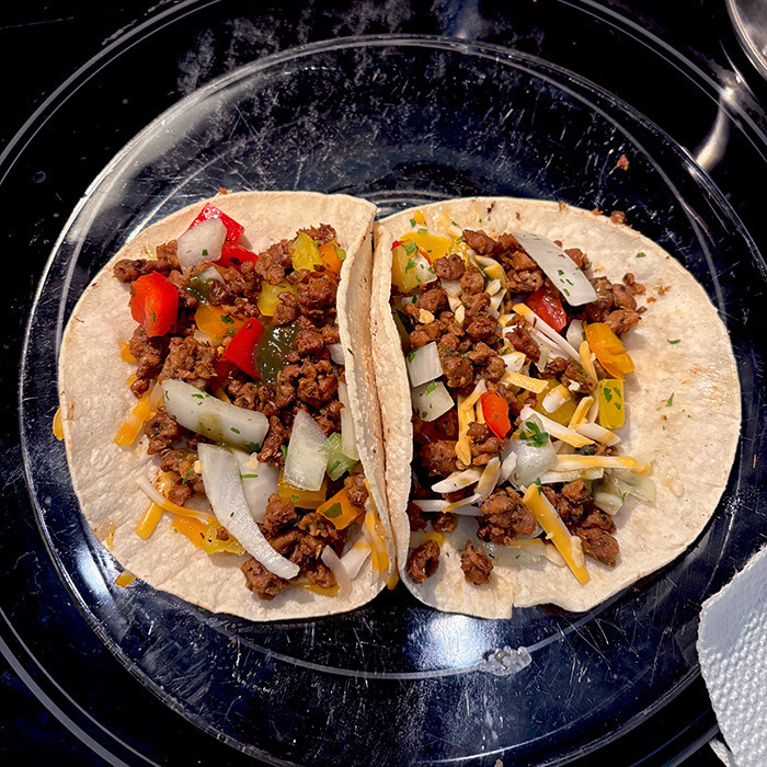 Two tacos filled with vegan ground meat (chorizo), diced vegetables (pico de gallo), and vegan cheese are served on a clear glass plate.