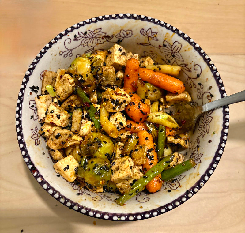 Auto-generated description: A bowl of stir-fried tofu with carrots, Brussels sprouts, and green beans, garnished with black sesame seeds.
