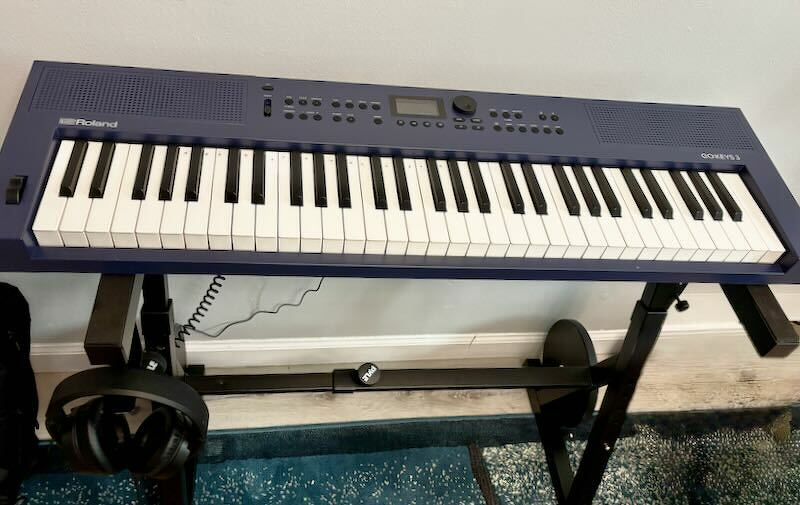 A blue Roland GO:KEYS 3 keyboard rests on a stand with headphones hanging below it. A barbell under one of the beams, and a subpar AI cleaning technique can be seen on the carpet at the bottom right, where JTR removed his used socks from the frame. Tsk tsk.