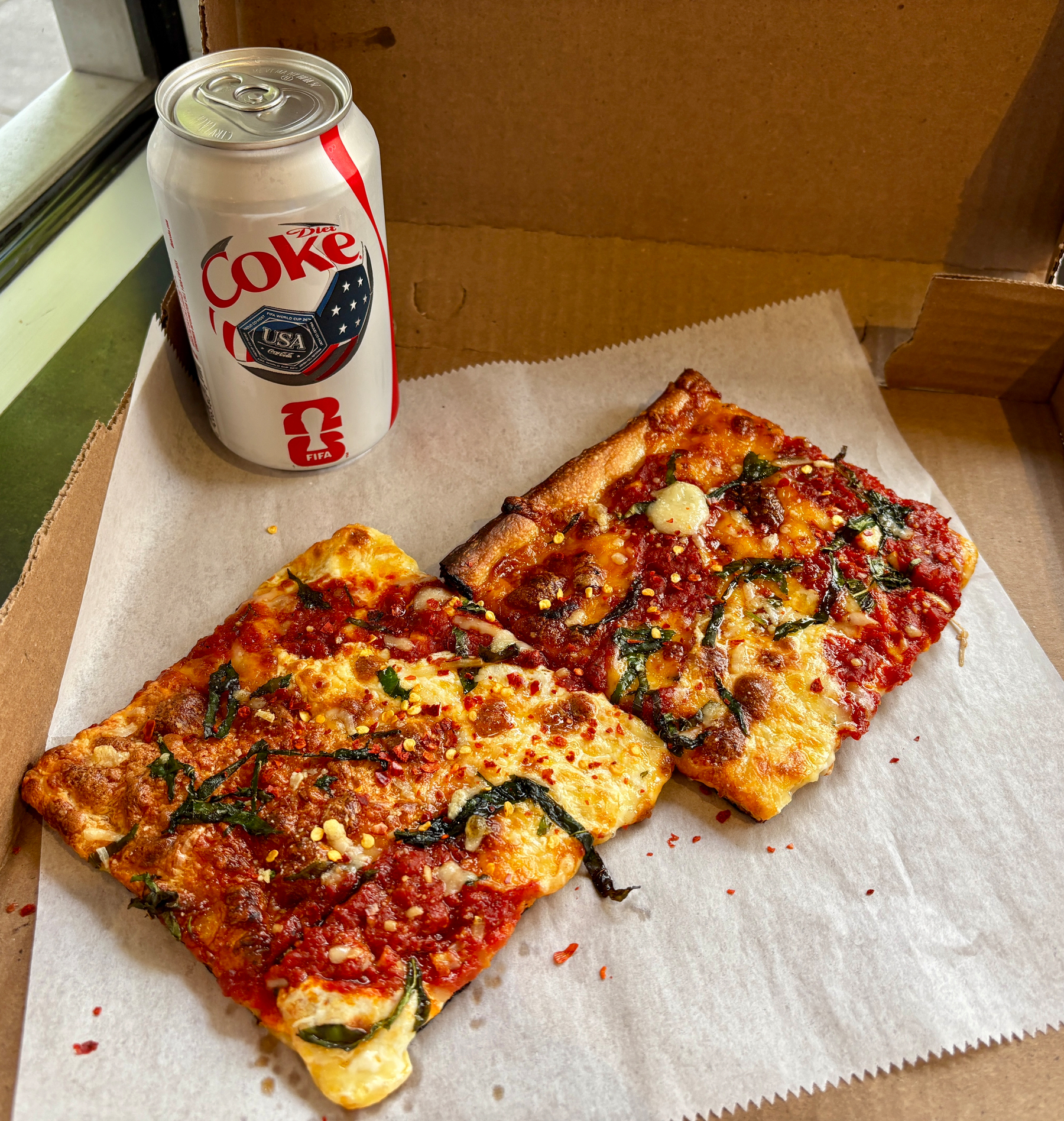 Two slices of pizza with spinach and cheese are on a cardboard box next to a can of Diet Coke.