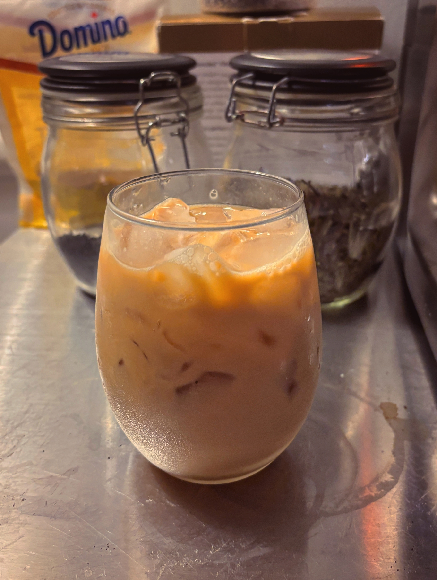 A glass of iced coffee or iced latte sits on a metal surface with jars and a bag of Domino sugar in the background.