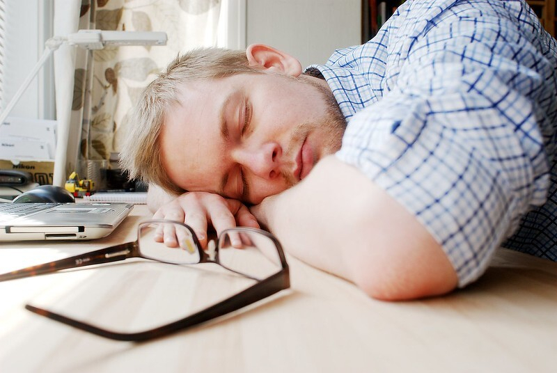 Auto-generated description: A man in a checkered shirt is sleeping on a desk next to a pair of glasses and an open laptop.