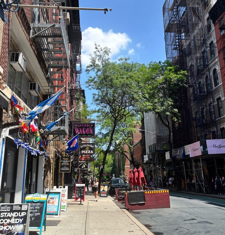 Auto-generated description: A lively urban street scene features colorful flags, restaurant signs, outdoor seating, and pedestrians under a bright blue sky with trees lining the sidewalk.