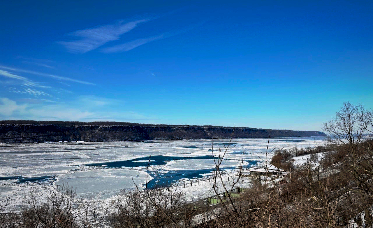 A frozen river stretches across the landscape surrounded by bare trees under a clear blue sky.