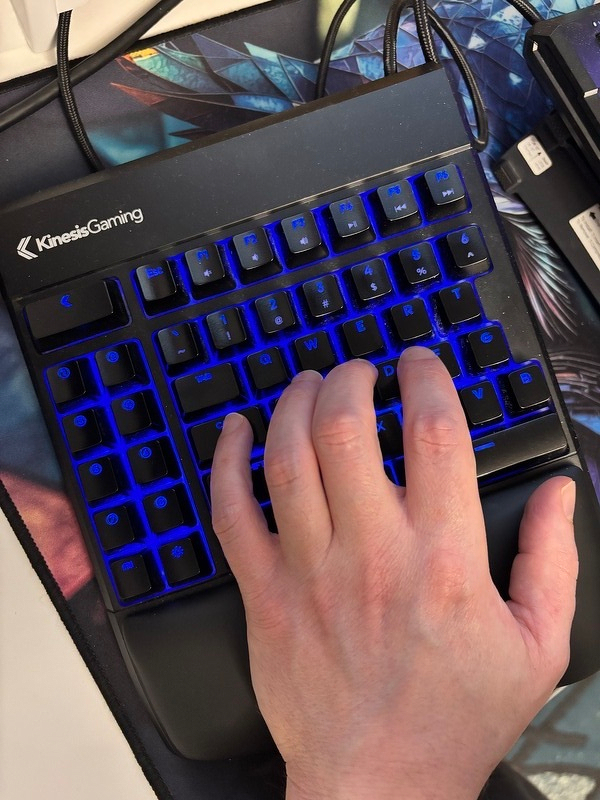 A hand is positioned on the left side of a  Kinesis Gaming mechanical keyboard with blue backlit keys.