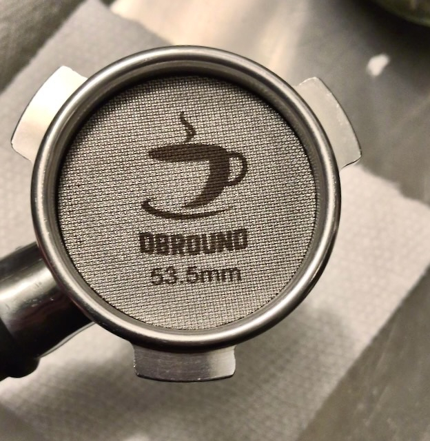 A coffee tamper is shown with a coffee screen inside with the text DBROUND 53.5mm on its surface.