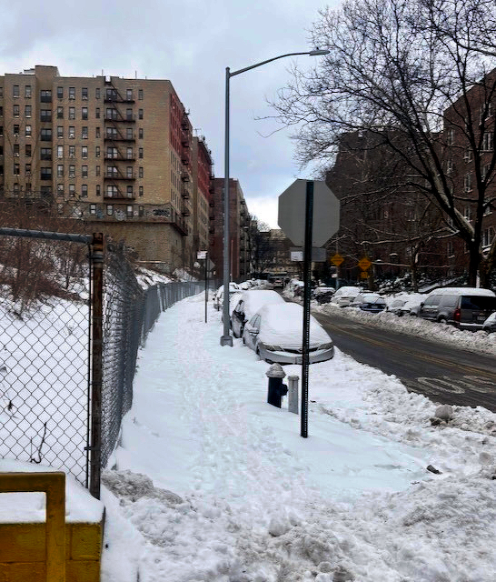 A snowy urban street scene with parked cars covered in snow, a snow-covered sidewalk, and a backdrop of apartment buildings. There’s snow everywhere, OK? Lots of snow.