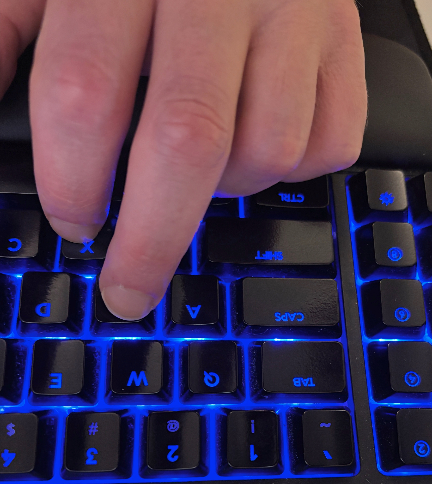 A hand is resting the keys X and A with the little finger curled over the Ctrl key on a backlit keyboard with blue lighting.