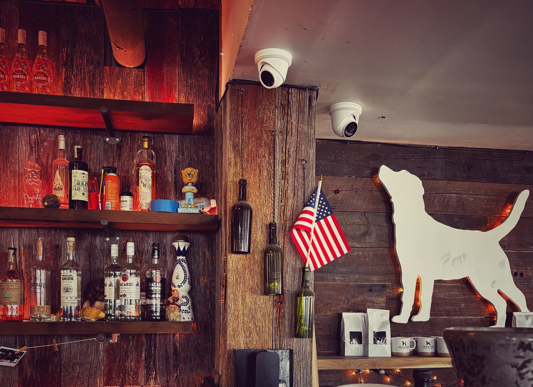 A cozy bar interior features shelves with various liquor bottles, a large white dog silhouette decoration, and mounted security cameras above an American flag.
