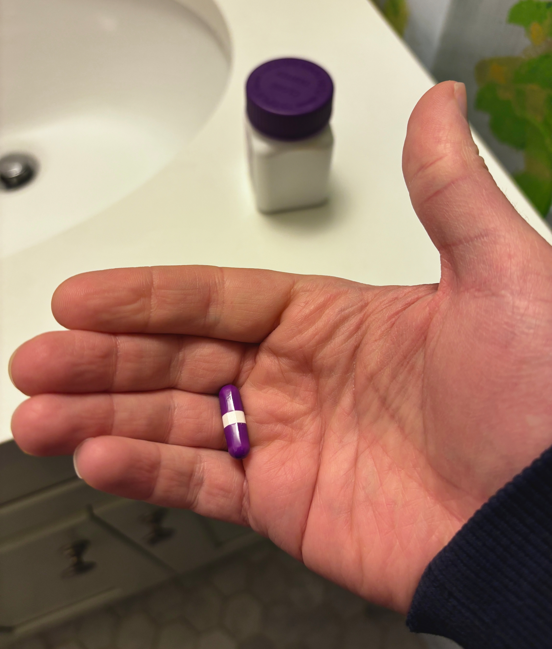 A hand holds a purple capsule pill over a bathroom counter with a bottle in the background.