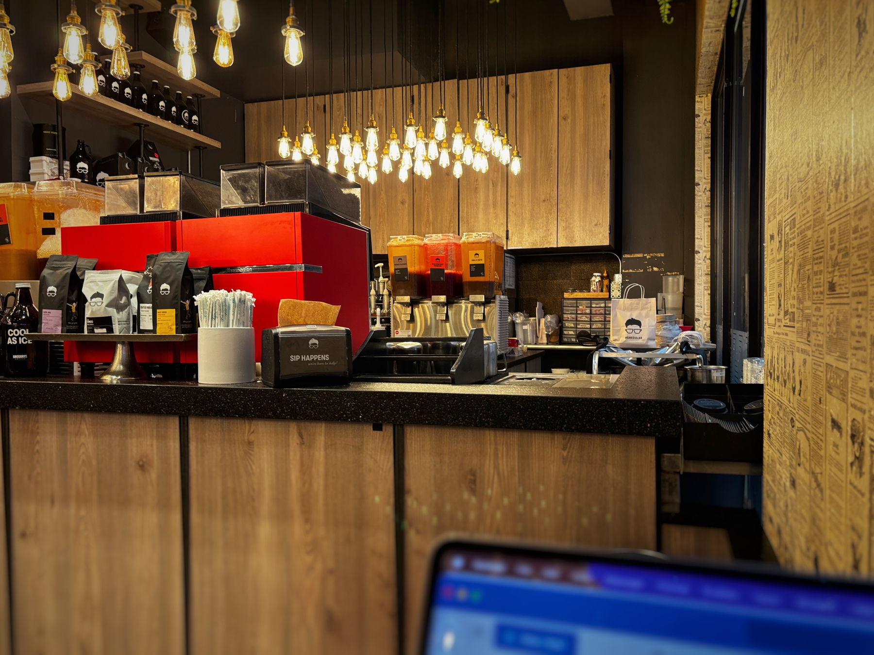 Auto-generated description: A cozy coffee shop counter features hanging light bulbs, red coffee machines, and various coffee supplies.