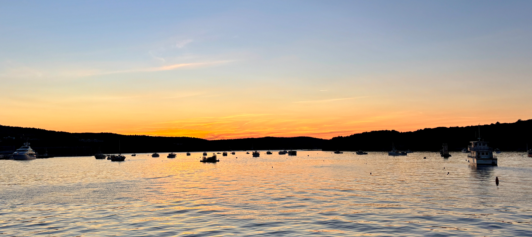 A calm body of water with numerous boats anchored and a vibrant sunset sky in the background.
