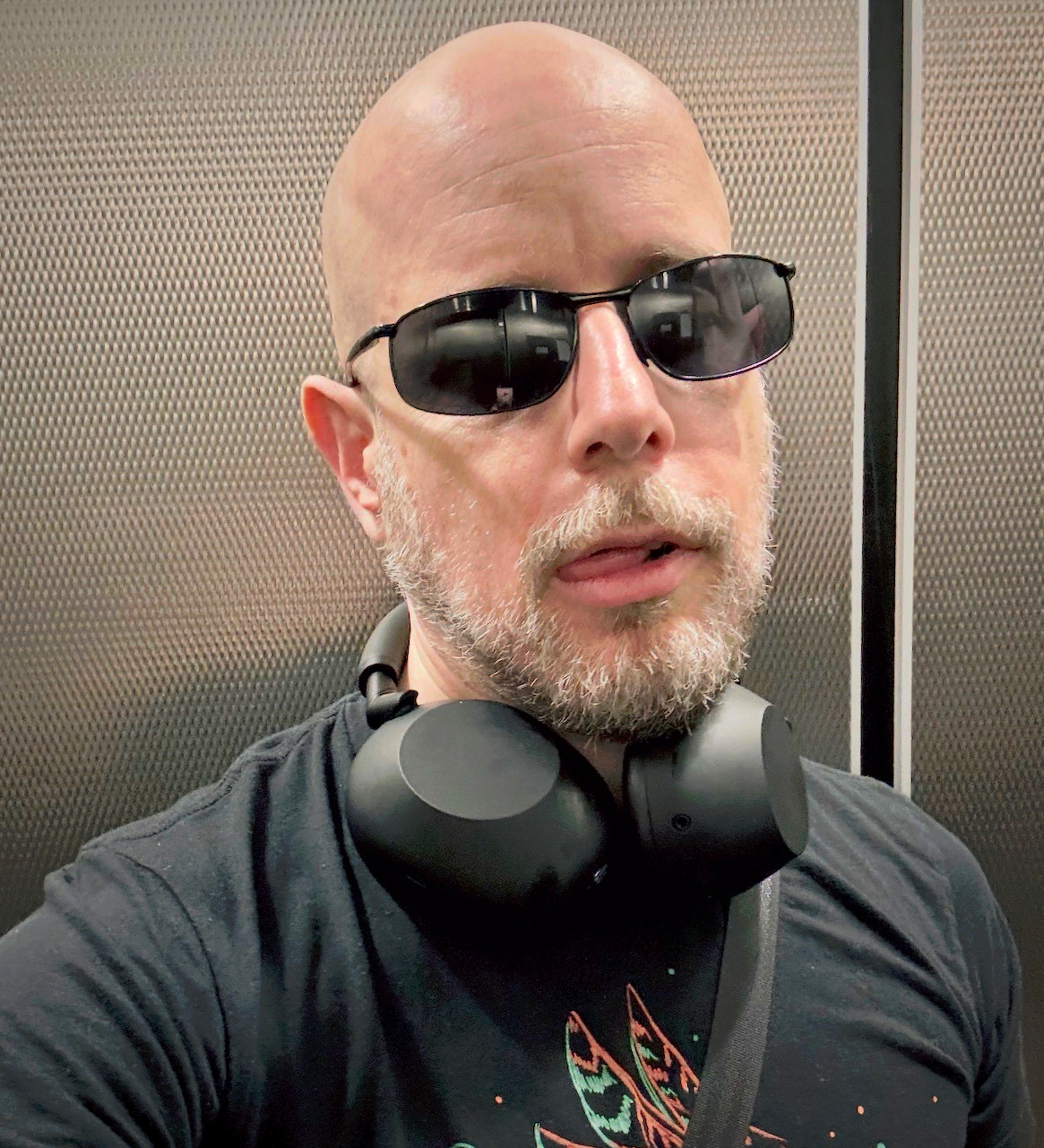 A bald man with a beard is wearing dark sunglasses, a black t-shirt with a graphic design, and large black headphones around his neck.